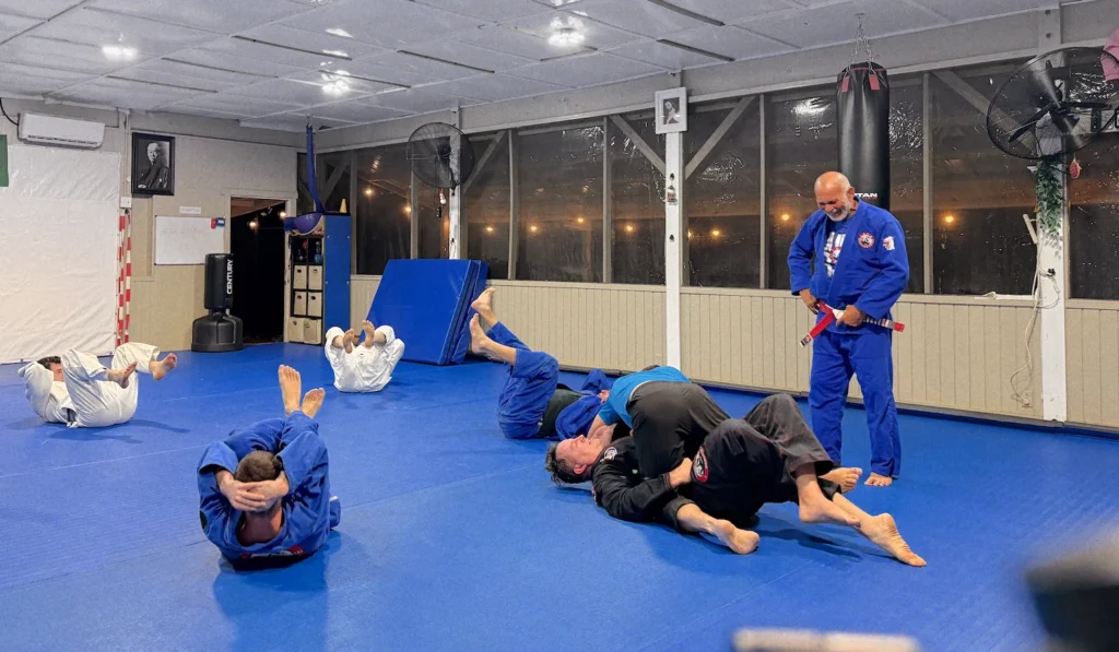 Adult BJJ students doing floor cooldown exercises on blue mats with instructor watching, Team Carvalho dojo flags visible