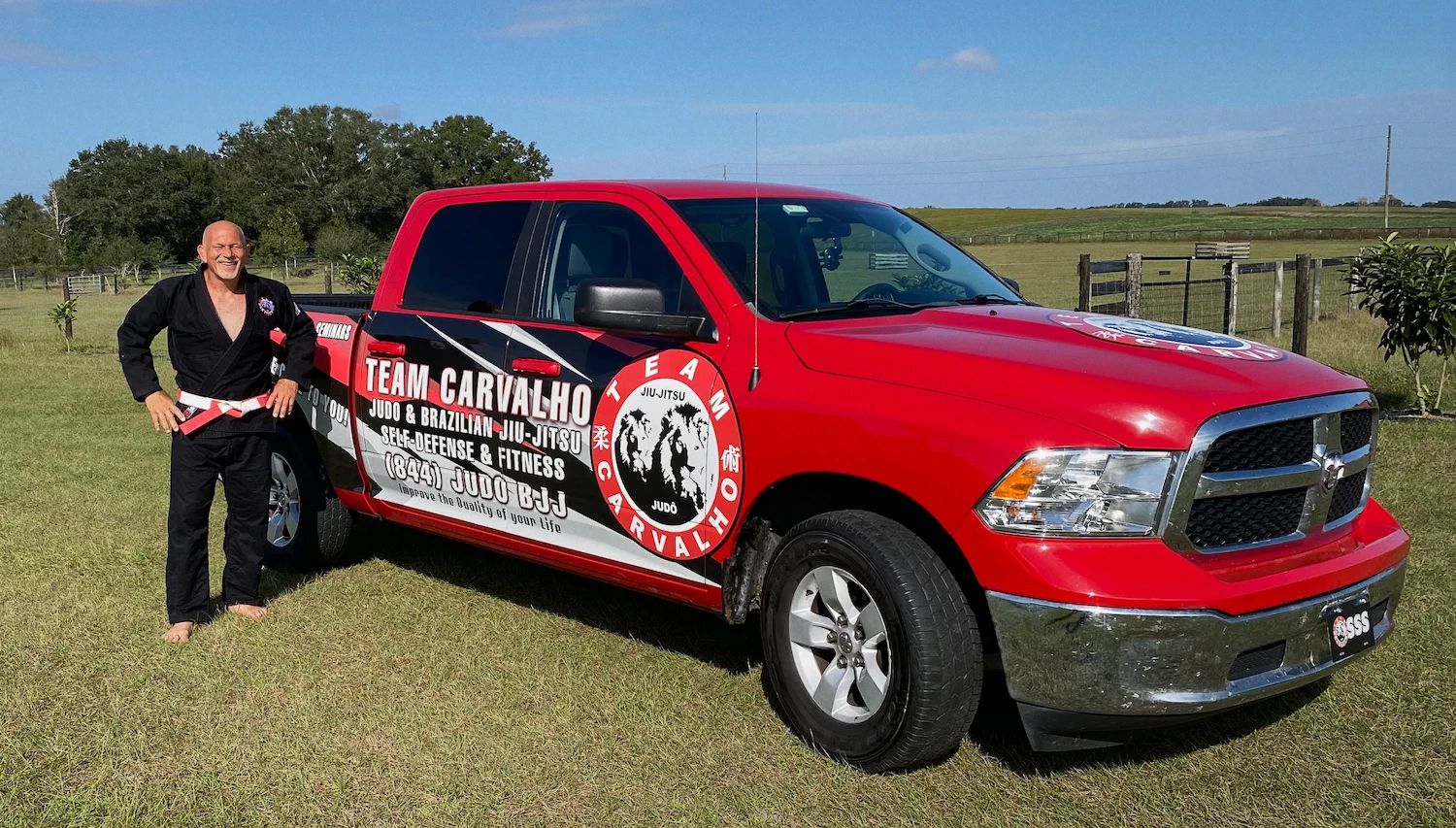 Team Carvalho BJJ instructor in black gi standing beside the driver side of the Team Carvalho branded red TCMS pickup truck