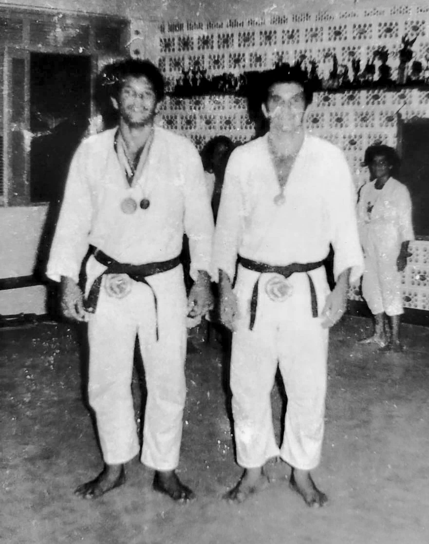Vintage black-and-white photo of two young male Judo practitioners in white gis with black belts and competition medals inside a dojo